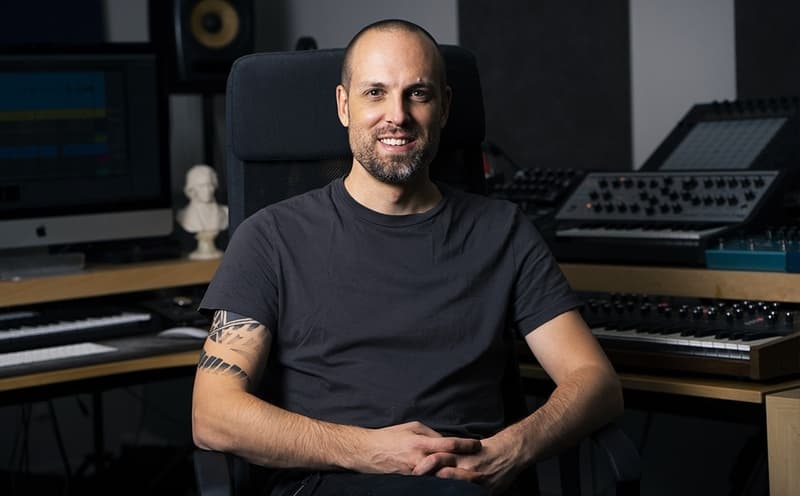 Tim Penner seated in his studio, smiling, with monitors and synth visible behind